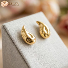 Gold earrings on a gray fabric surface with 'ZZILYA' branding.