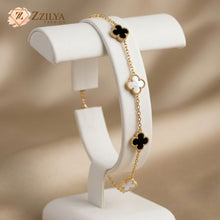 A delicate gold chain bracelet featuring alternating black and white four-leaf clover charms. The bracelet is displayed elegantly on a white curved jewelry stand. The ZZILYA Fashion logo is visible in the upper-left corner against a soft, draped beige background.