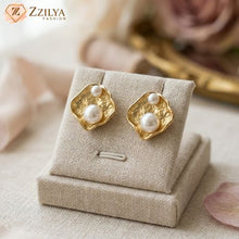 ZZILYA Fashion gold-tone anti-tarnish statement stud earrings featuring an organic hammered texture and two asymmetrical white pearls, displayed on a beige linen jewelry stand.