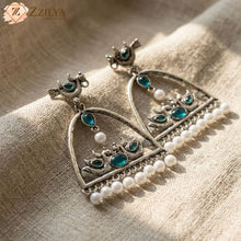 Close-up product shot of Zzilya Fashion's trendy silver oxidized earrings featuring an arch shape decorated with traditional peacock and floral motifs, inlaid with deep teal stones, and finished with a neat row of white hanging pearls resting on a textured linen background.