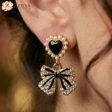 Close-up of an earring with a bow design and heart-shaped gem, worn by a person.