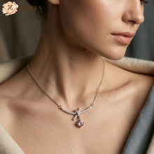 A woman wearing a delicate silver necklace by ZZILYA Fashion featuring a pavé-encrusted bow design and a dangling round pink gemstone pendant.