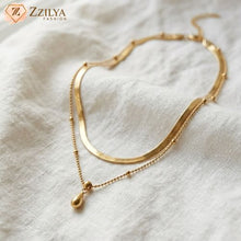 ZZILYA Fashion double-layered gold neckchain featuring a flat herringbone chain and a beaded chain with a polished teardrop pendant on a white fabric background.