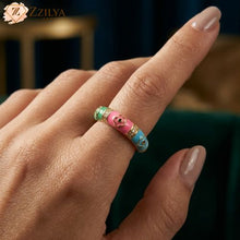 A golden anti-tarnish band ring by Zzilya Fashion featuring pink, blue, and green enamel segments with gold flower designs and sparkling zircon accents, worn on a model's hand.
