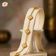 A delicate gold chain bracelet featuring gold four-leaf clover charms. The bracelet is displayed elegantly on a white curved jewelry stand. The ZZILYA Fashion logo is visible in the upper-left corner against a soft, draped beige background.