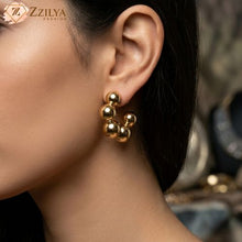 A close-up photograph of a woman wearing a golden beaded hoop earring. The earring is a small hoop with multiple polished gold beads of varying sizes clustered together. The woman has dark hair styled in an updo and her profile is visible. She has a subtle and stylish look. The background is slightly blurred and features warm, golden tones. The 