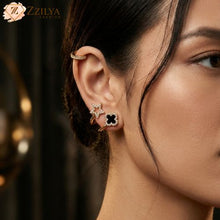 A close-up product photo of a pair of elegant, gold wrap-around earrings. One part of the earring features a black quatrefoil (clover) design, and the other part is a delicate, pavé-set shimmering star that sits below the earlobe when worn. The earrings are displayed on a light velvet holder. The text 