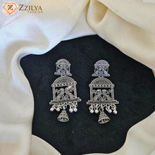 A pair of silver-toned traditional Indian earrings with intricate filigree and bird motifs, displayed on a black textured surface with draped cream fabric.