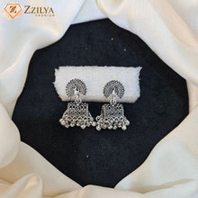 A pair of silver-toned traditional Indian jhumka earrings with intricate filigree work and hanging bead chains, displayed on a white textured stand over a black felt surface with draped white fabric around it. The brand logo 'Zzilya Fashion' is visible in the top left corner.