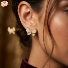Close-up macro photograph of a textured golden bow-shaped earring with a shimmering stone-studded center, displayed on a plush dark velvet background. Includes a branding tag for Zzilya Fashion with product details.