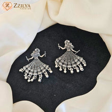 A pair of silver-toned Jhumka earrings with detailed filigree work and dangling bell charms, displayed on a black textured surface surrounded by draped cream fabric. The Zzilya Fashion logo is visible in the top left corner.
