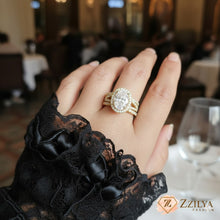 Close-up of a luxury oval-cut diamond halo ring with a triple-row gold pavé band worn on a hand with a black lace sleeve, set against a blurred high-end restaurant background.